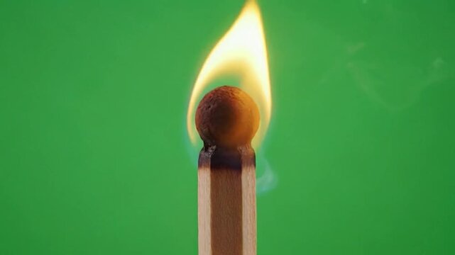 A close-up shot of a burning matchstick with a bright yellow flame against a solid green background