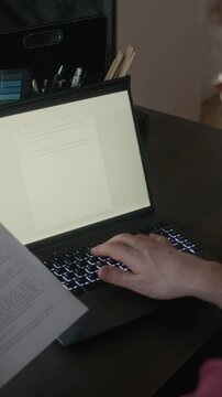 Vertical high angle view of mature Caucasian male author holding paper with short story draft, writing clean copy on laptop