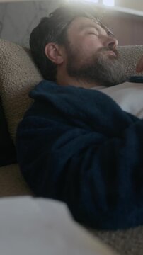 Vertical of bearded Caucasian male author taking nap on couch in living room while working on new novel