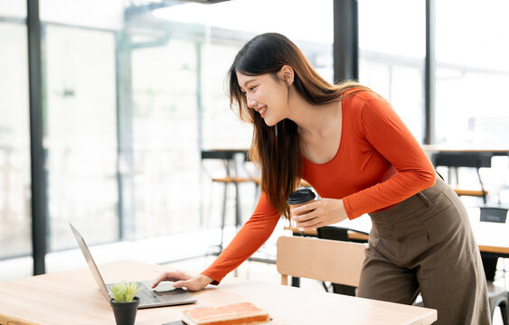 Cheerful Asian woman freelancer using laptop in cafe while holding coffee.