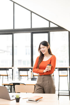 Happy Asian woman freelancer holding coffee while working in modern coworking space.