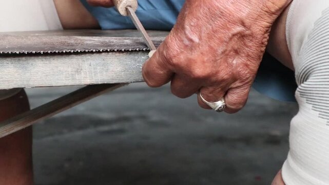 The hands of an Asian person are sharpening a hand saw with a metal file, honing the saw teeth.