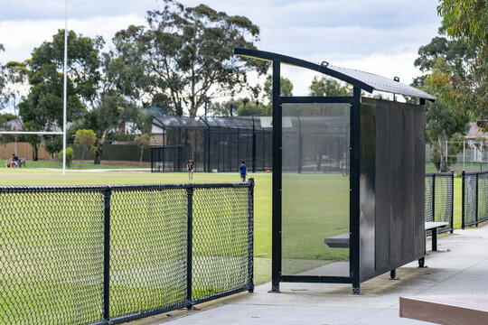 A metal shelter or sports dugout at the edge of a suburban recreational reserve, overlooking a green sports field with goal posts and children playing in the background. Melbourne Australia.