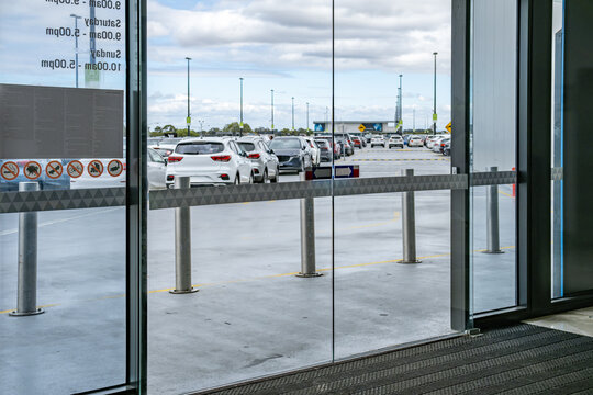 Automatic sliding glass doors of a shopping center, overlooking a large outdoor parking lot filled with cars. The entrance features safety decals, opening hour signage, and metal bollards 