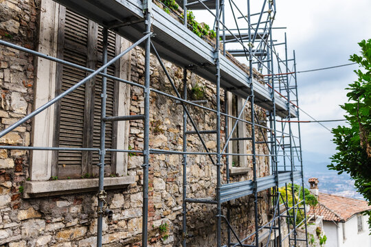 Historic stone building facade with wooden shutters covered by metal scaffolding during renovation in Italy
