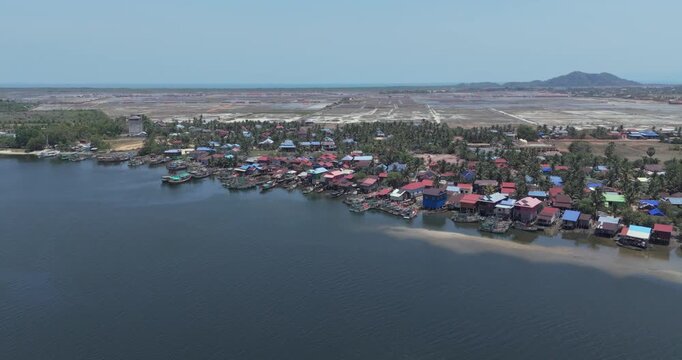 Forward aerial view of a coastal fishing village with colorful rooftops and moored boats, bordering expansive salt pans under a clear sky.