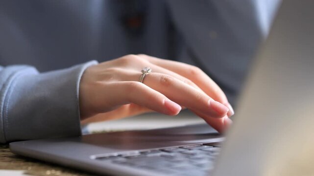 Close-up of a person's hands typing on a laptop. Fingers press keys with focus on ring on left hand. Blurred background shows gray sleeve and wooden surface. Neutral lighting highlights hand