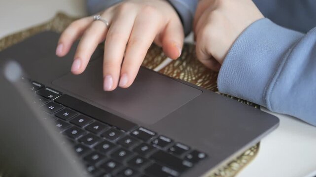 Close-up of hands using laptop touchpad. Fingers scroll and tap on dark gray surface. Person wears light blue sleeves. Ring visible on left ring finger