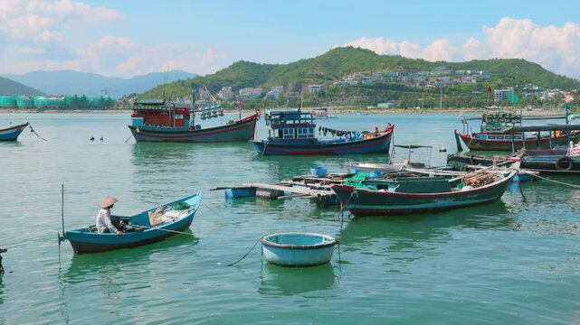 A lone fisherman in a conical hat rows a small boat in Hon Meiu Island, Nha Trang, Vietnam, surrounded by larger fishing vessels in the bay.
