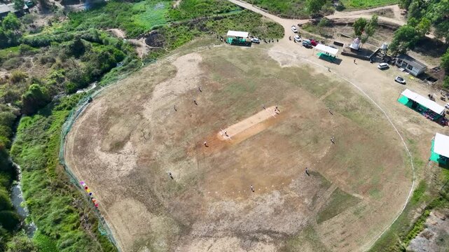 An aerial view captures a rustic cricket ground set amidst a natural landscape, showcasing a local match in progress. The circular field is uneven and dry, with patches of grass and exposed soil.
