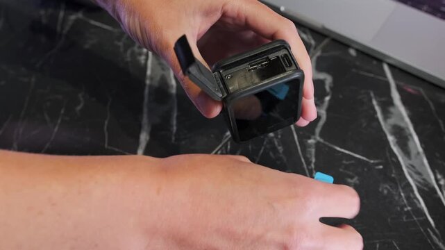 Close-up of hands inserting batteries into action camera compartment with clicking lock mechanism, preparing device for recording and power supply on workspace desk. Inserting action camera batteries