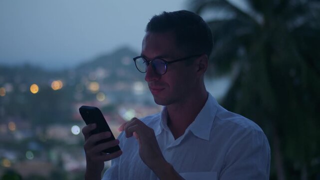 Internet addiction concept as man scrolls smartphone at dusk with screen reflected in glasses. Excessive screen time and constant online browsing highlight digital dependency lifestyle.
