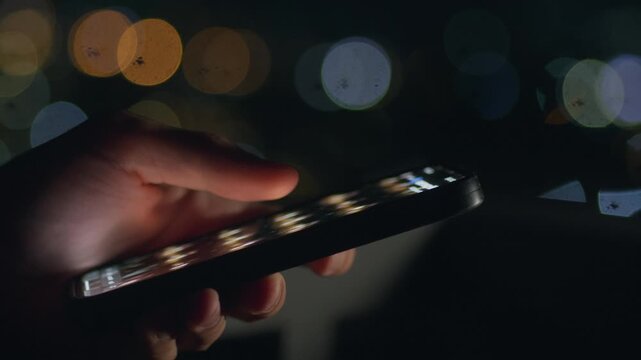 Hand holding smartphone at night while scrolling screen with city bokeh lights. Night smartphone use shows modern urban lifestyle night technology mobile habits and late night digital activity.