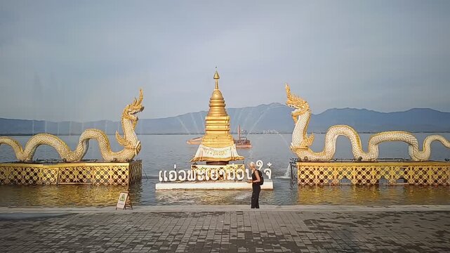 A traveler photographing the iconic Naga sculptures and Buddhist stupa at the waterfront of Kwan Phayao, Northern Thailand.