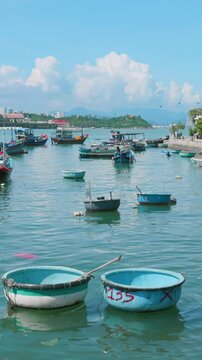 A scenic view of traditional Vietnamese coracle boats and fishing vessels moored in the turquoise waters of Hon Meiu Island, Nha Trang, Vietnam, with coastal buildings in the background.