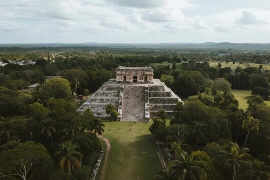Aerial view of an ancient Mayan pyramid surrounded by lush green forest and open grassland under a cloudy sky