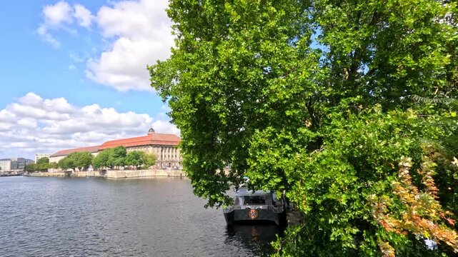 View of historic building on spree river bank in Berlin with lush green tree and boat in foreground.