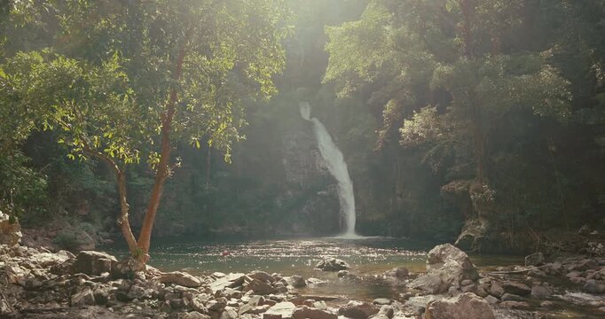 Tropical waterfall flows into natural pool surrounded by lush green trees and rocks. Sunlight filters through jungle canopy creating bright flare and serene atmosphere.