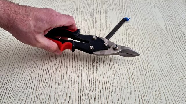 Hand holding red and black metal snips above light wooden surface near small metal strip with blue end, highlighting cutting tools, precision handling, and repair essentials