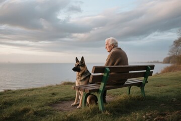 Fototapeta premium Elderly person sitting on a bench with a German Shepherd dog by the water at dusk