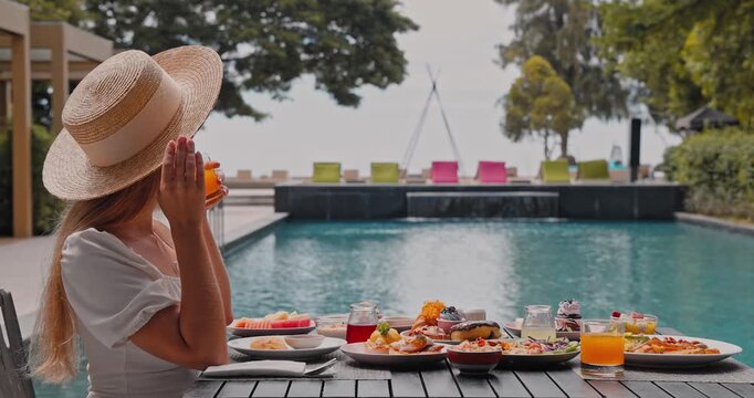Woman wearing straw hat drinking orange juice during breakfast, luxury tropical resort poolside setting, summer vacation, high-end travel marketing background