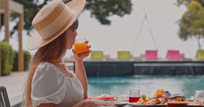 Woman wearing straw hat drinking orange juice, luxury tropical resort poolside breakfast table with pastries and fruit, summer vacation travel advertising