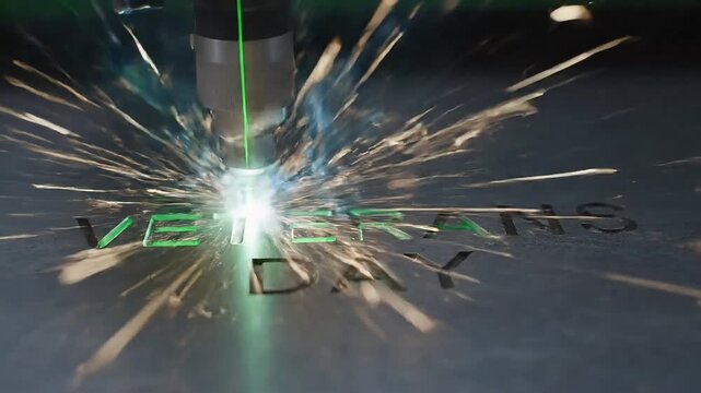 Laser engraving machine carving Veterans Day text into dark metal plate creating bright sparks and smoke in this high quality industrial motion background