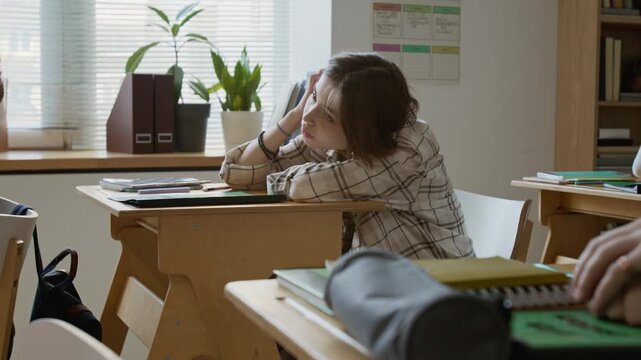 Medium side shot of distracted, apathetic teenage Caucasian girl lying on desk and playing with hair during lesson at high school, expressing lack of interest, motivation and boredom