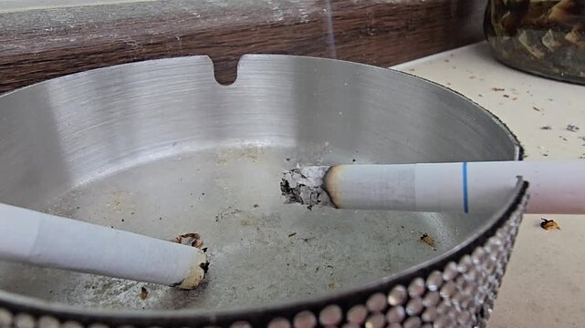 Close up of burning cigarette with ash in metal ashtray on table, smoking concept, unhealthy lifestyle and bad habit 
