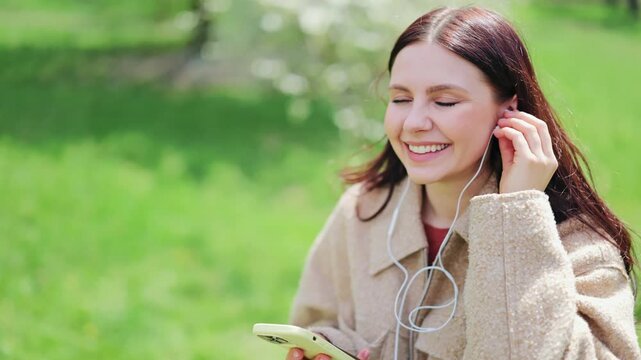 Young woman smiles, captivated by music through earphones, enjoying peaceful moment outdoors amidst vibrant green park. Pure joy evident on face.