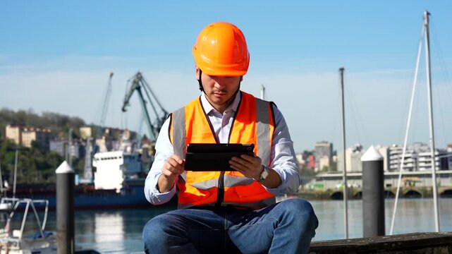 Young asian male engineer in a hard hat and safety vest sitting at the commercial dock, using a digital tablet for managing logistics and container ship operations with a smile