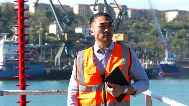 Professional asian cargo port engineer wearing a safety vest using a digital tablet for logistics management and quality control, then looking towards the camera with a determined expression