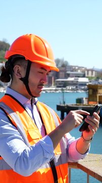 Young asian male engineer in a hard hat and safety vest using a smartphone for logistics management at a commercial dock, with cargo cranes and the seaport visible in the background