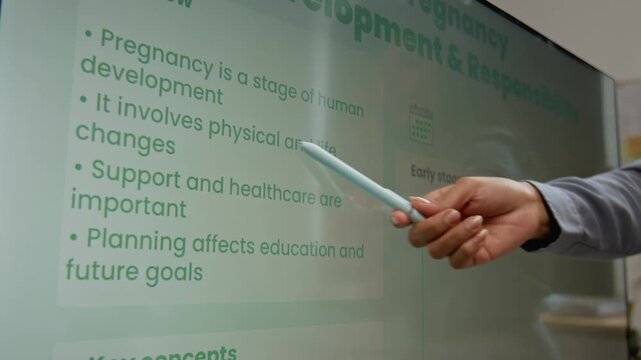 Medium close-up of hand of unrecognizable female teacher pointing to slide on screen during lesson on pregnancy in high school, highlighting main concepts of decision making and responsibility