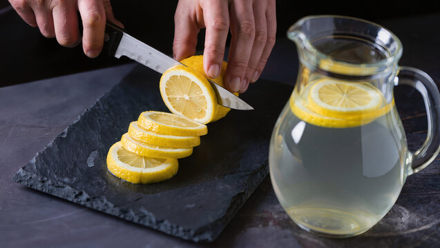 Hands Slicing Fresh Lemon Halves on Slate Board Next to Pitcher of Lemonade