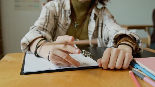 Medium midsection shot of unrecognizable young teenage female high school student in casual outfit fidgeting with pencil while writing test at lesson, with doubt and lack of comprehension