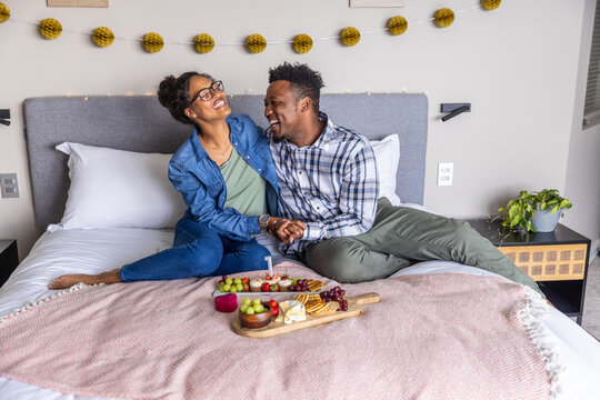 Romantic couple laughing and holding hands on bed with wooden board of grapes cheese crackers