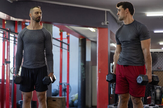Holding hex dumbbells, two fit men training at gym near red rig, plyo box, medicine balls