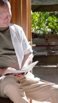 Vertical video: Reading senior man in white shirt at door turning book pages to relax in sunlight