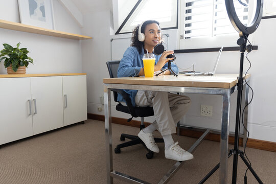 Adult non-binary podcaster wearing white headphones speaking into condenser microphone at home desk
