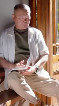 Vertical video: Sunlight drawing elder on bench by door reading book and glancing outside white top