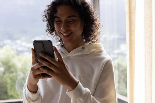 Non-binary adult standing by large window holding smartphone with hands, wearing hoodie and rings