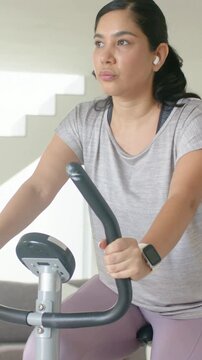 Vertical video: Gripping bars, woman in grey top pedaling bike at home wearing earbuds for exercise