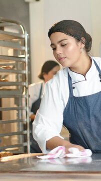 Vertical video: Wiping chef in coat  and  dark apron cleaning bakery counter with pink cloth post tr