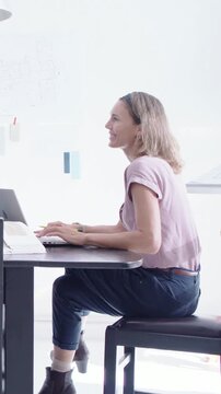 Vertical video: Typing woman in pink top reaching, peer appearing, placing notes on glass by laptop