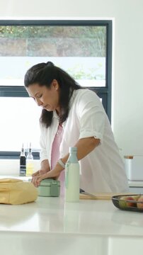 Vertical video: Packing lunch, woman in shirt closing green box at home near yellow bag and bottle