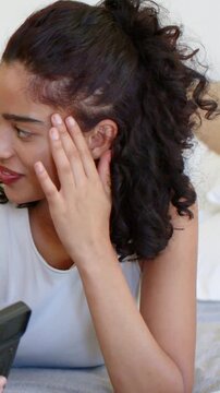 Vertical video: Glancing woman in white tank top sitting up on bed in bedroom, reacting to phone