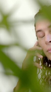 Vertical video: Framing plant blur showing person taking call holding phone at home in light top