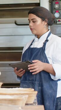 Vertical video: Checking tablet, baker in denim apron at bakery checking proofing baskets gesturing
