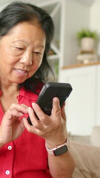 Vertical video: Using black phone, Asian woman in red top tapping and reading on couch, pink watch
